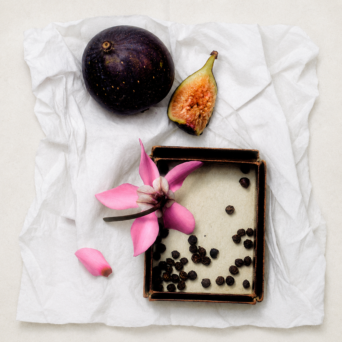 A styled flat-lay featuring a whole and halved purple fig, a pink flower, and small dark seeds arranged on a crumpled white paper sheet.